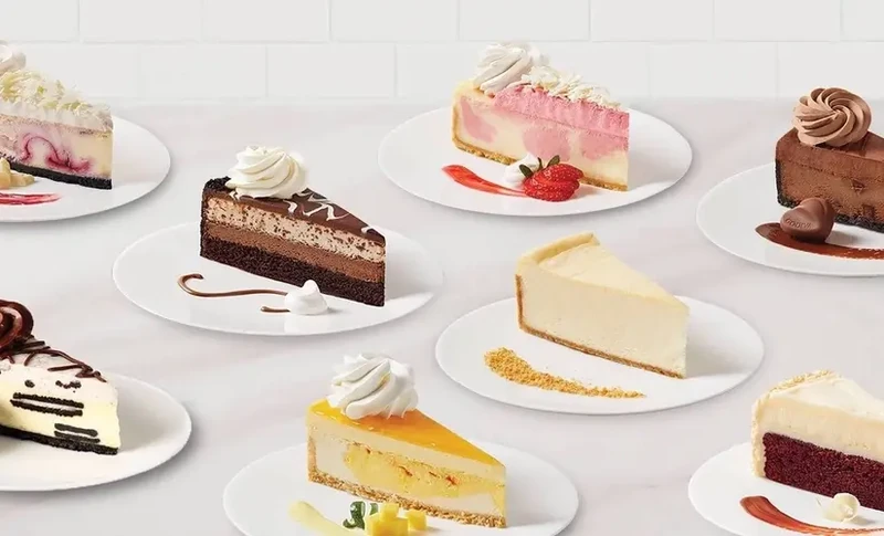 Assorted cheesecake slices on white plates, finished with whipped cream, fruit and chocolate
