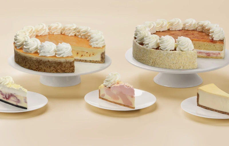 Assorted Cheesecake Factory Bakery cakes and cheesecake slices on stands and plates against a peach background