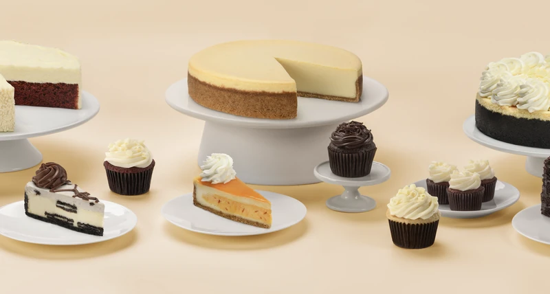 An assortment of whole cheesecakes, plated cake slices, and cupcakes arranged on stands against a warm peach background